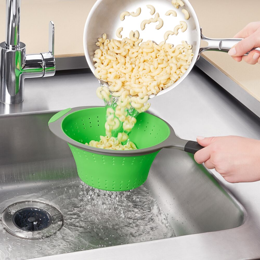 Hand using a green strainer to strain pasta into a metal sink. 