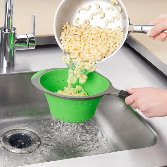 Hand using a green strainer to strain pasta into a metal sink. 