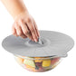 Gray silicone lid being placed on a bowl of fruit by a hand.