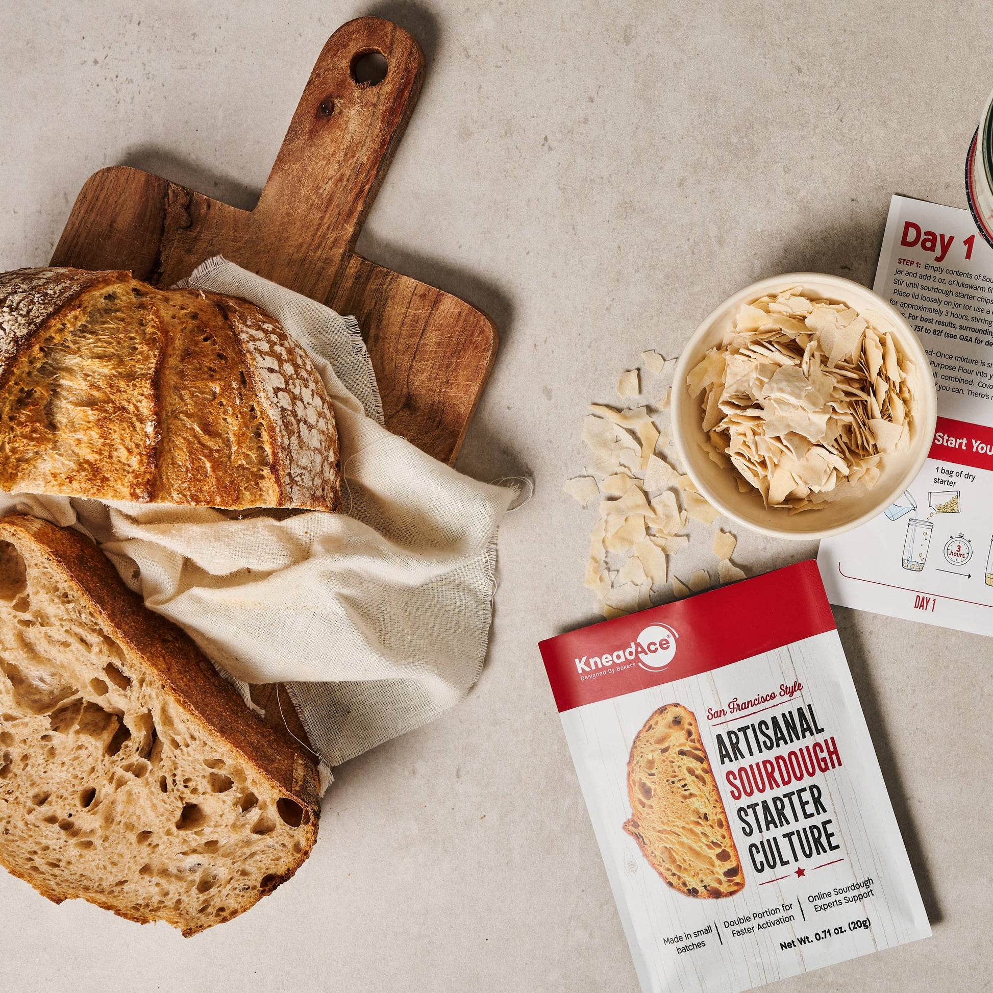 Bread with a wooden cutting board, sourdough starter culture package, and newspaper on a light gray surface.