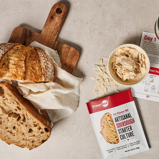 Bread with a wooden cutting board, sourdough starter culture package, and newspaper on a light gray surface.