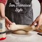 Person in a kitchen rolling dough with 'San Francisco Style' text on an apron.