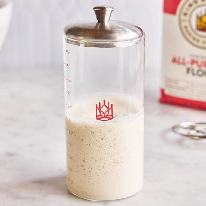 Glass Sourdough Storage Crock with a red logo, flour bag, and measuring spoon on a marble surface
