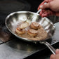 A stainless clad frying pan with a stay-cool handle, suitable for gas, electric, and induction cooktops frying fish in oil over a kitchen grill.