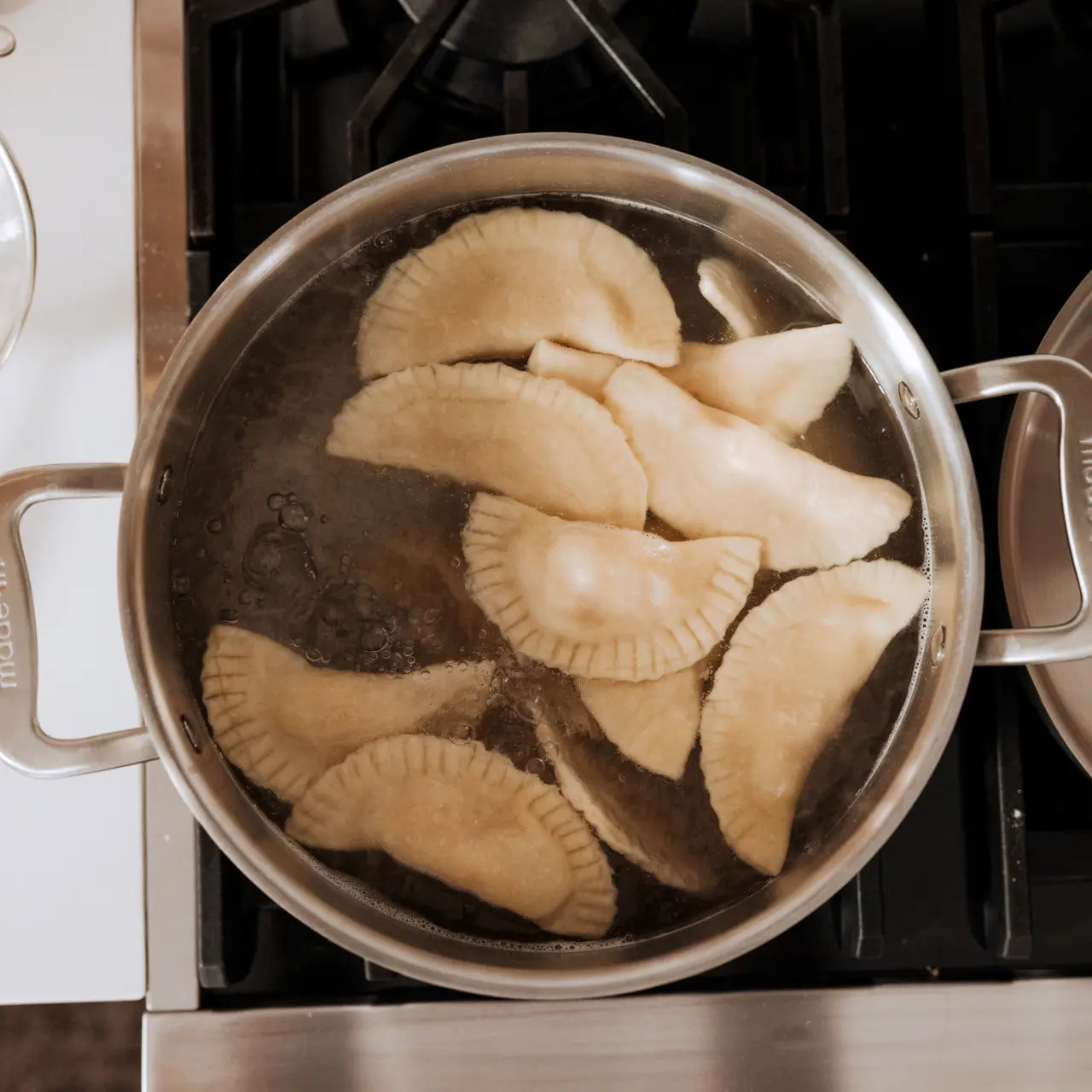 A silver stainless clad stock pot, featuring two handles boiling water with dough, on a kitchen stovetop.