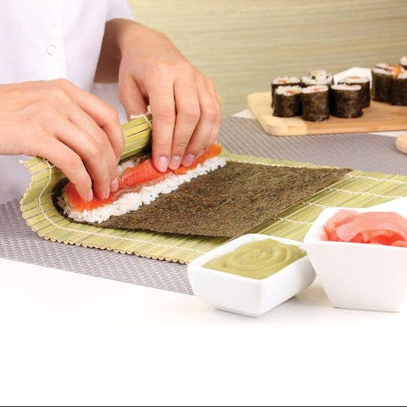Person making sushi with a rolling mat on a table with ingredients and a wooden board in the background.
