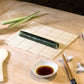 Sushi roll on a bamboo mat with cucumbers, soy sauce, and chopsticks on a wooden table.