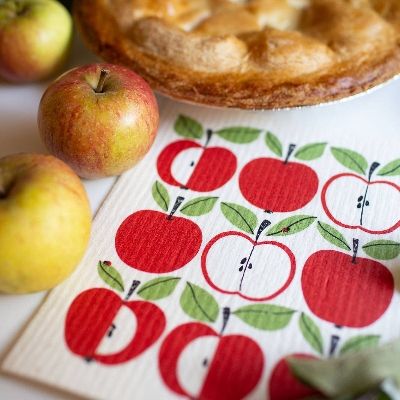 A close-up of a kitchen towel printed with a red and green apple pattern, styled with whole and sliced apples and a baked apple pie on a white surface.