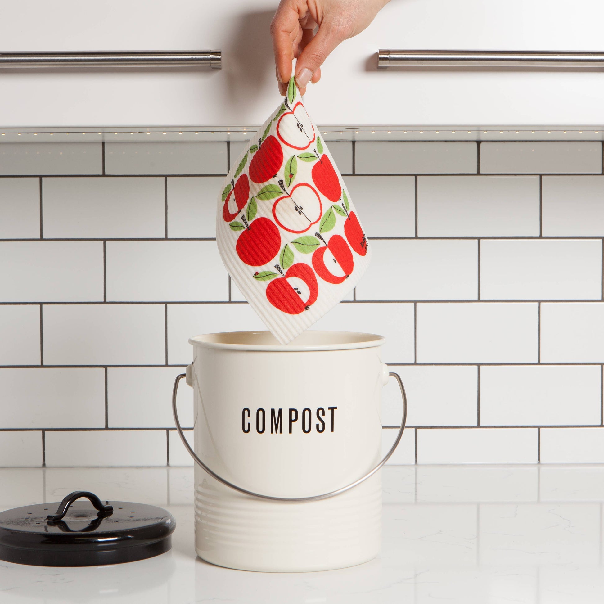 A hand holds a reusable cloth kitchen towel with a red apple and green leaf pattern above a white compost bin with a metal handle. The bin has the word COMPOST printed on the front, and a black lid sits on the counter to the left.