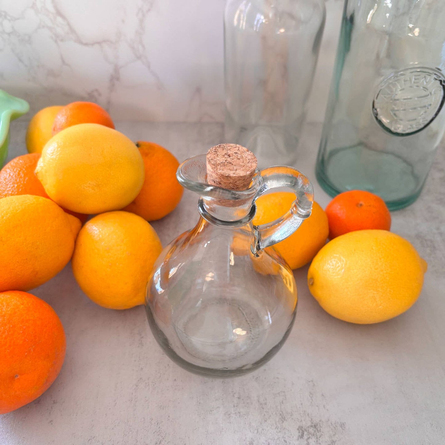 Clear glass bottle with cork stopper surrounded by oranges on a light surface