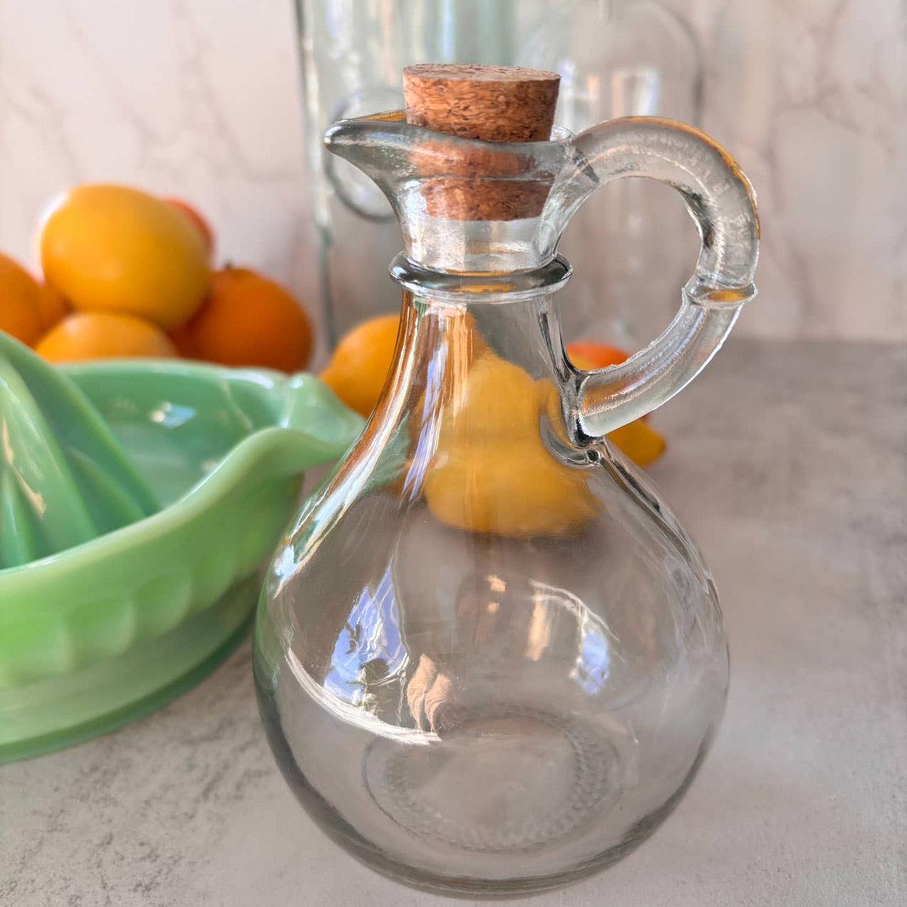 Clear glass carafe with cork stopper on a countertop with fruits in the background