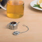 Tea infuser on a wooden surface with a glass of tea and a plate of lemon slices in the background.