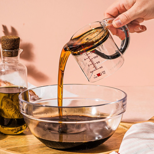 Person pouring liquid from a measuring cup into a glass bowl with a bottle of oil in the background.