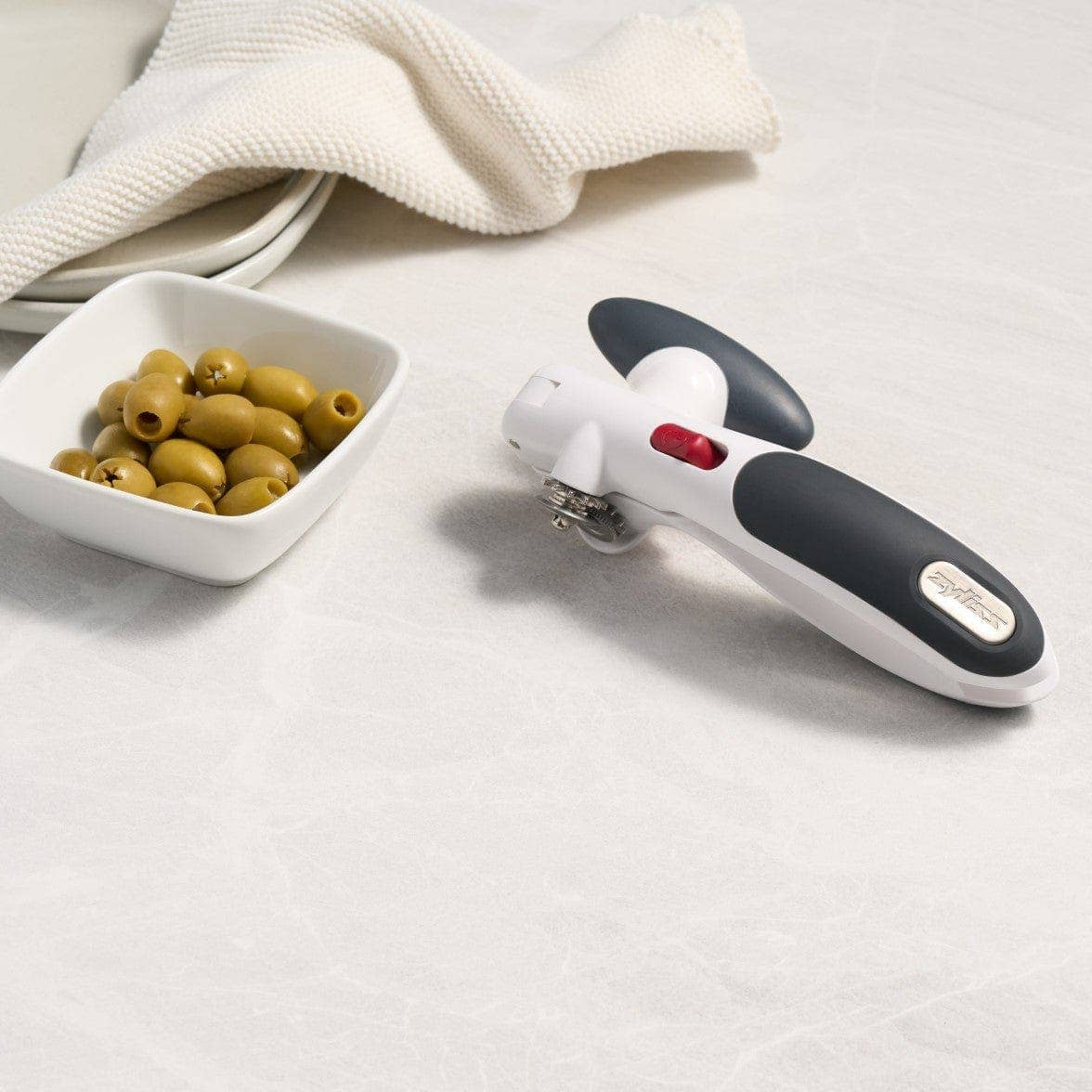 Handheld can opener with a small bowl of olives on a white surface