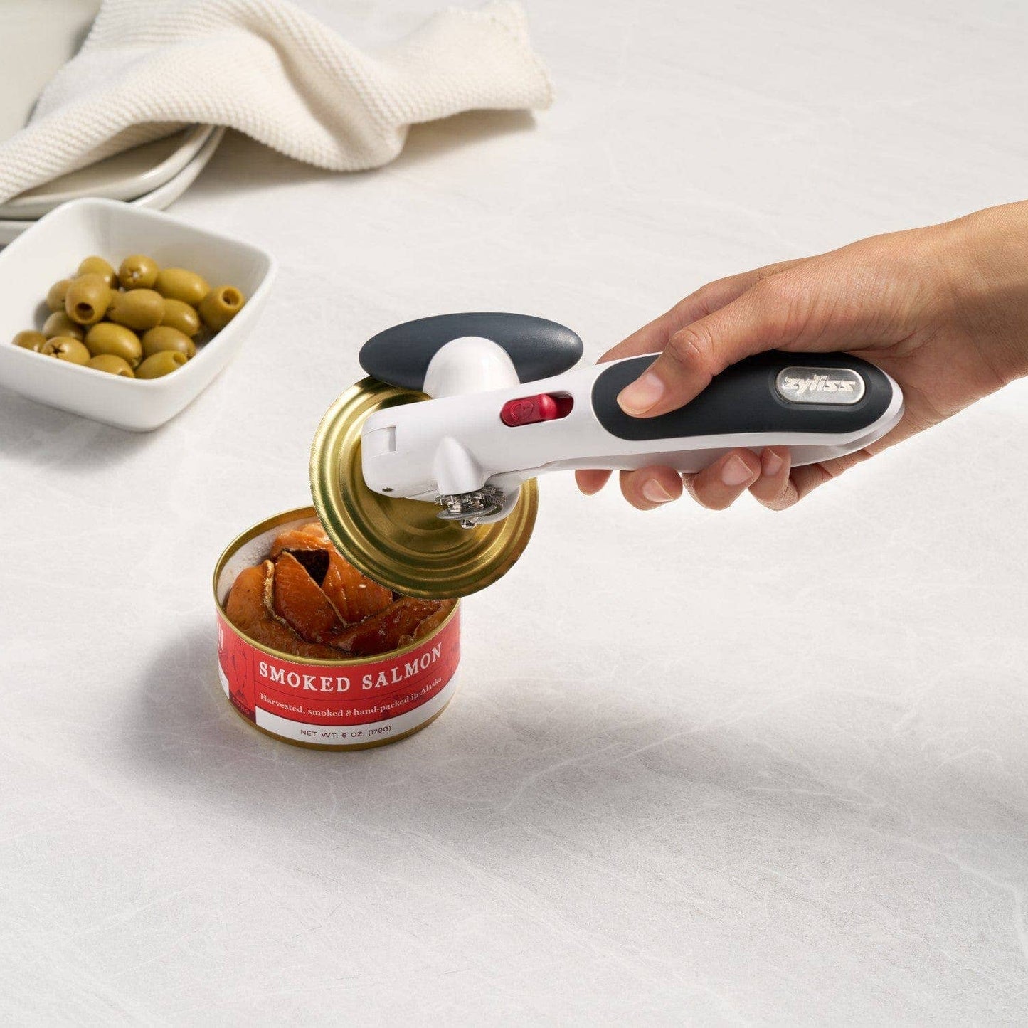Hand using a can opener on a can of smoked salmon with olives in the background.