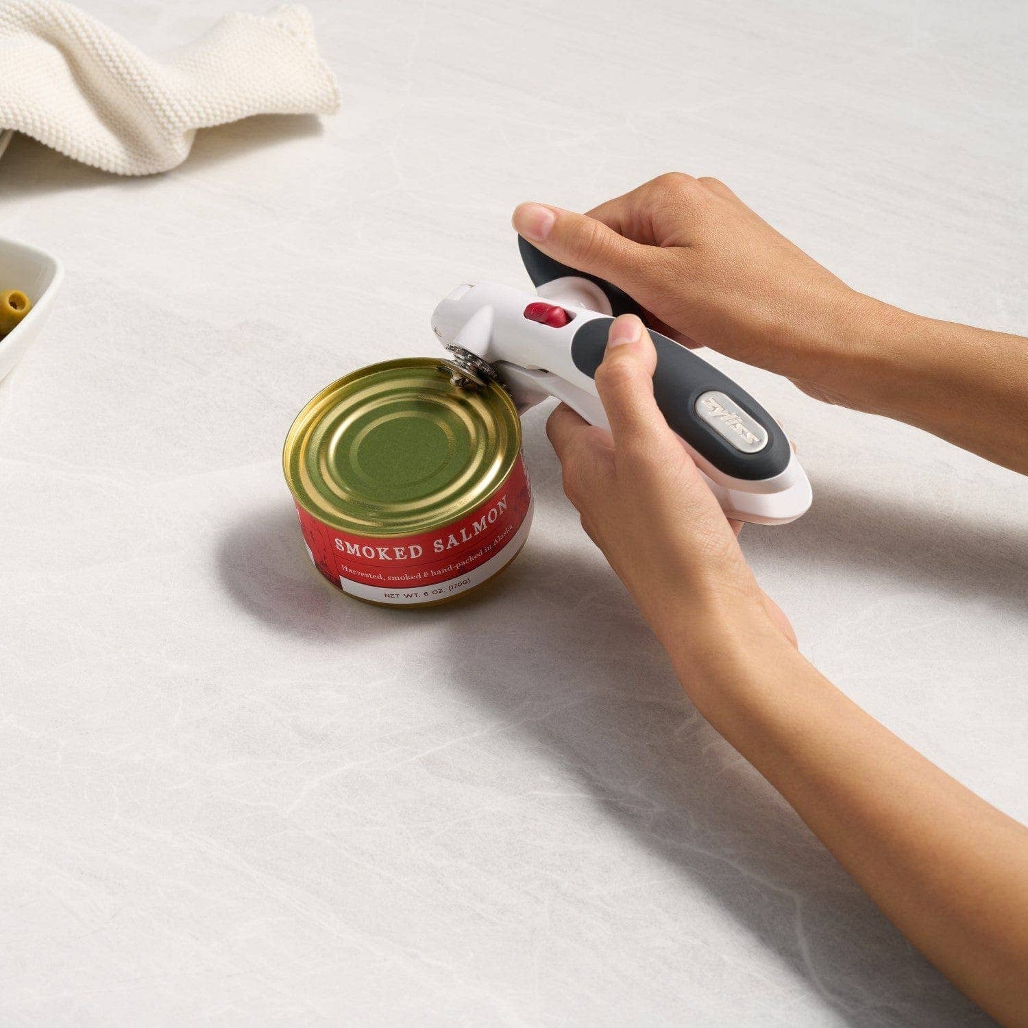 Person using a can opener on a can of smoked salmon with olives in the background.