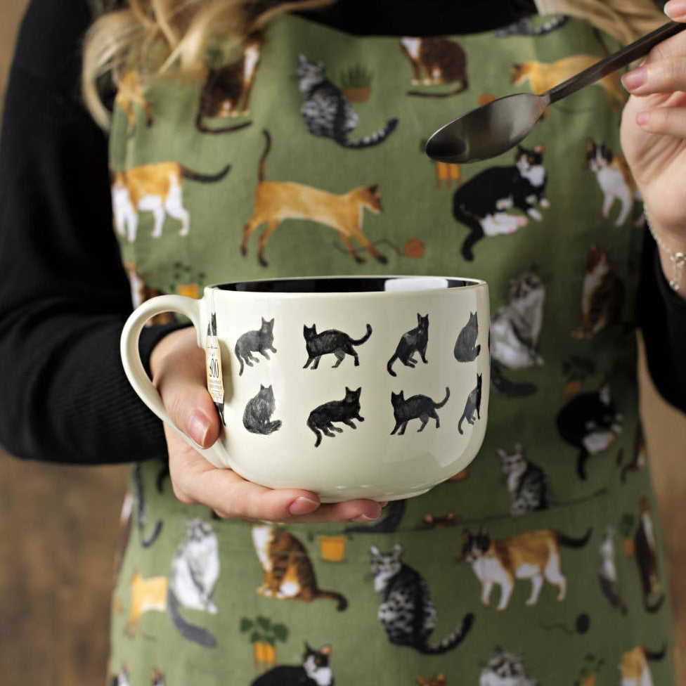 Person holding a mug with cat patterns and a spoon, wearing a green apron with cat designs.