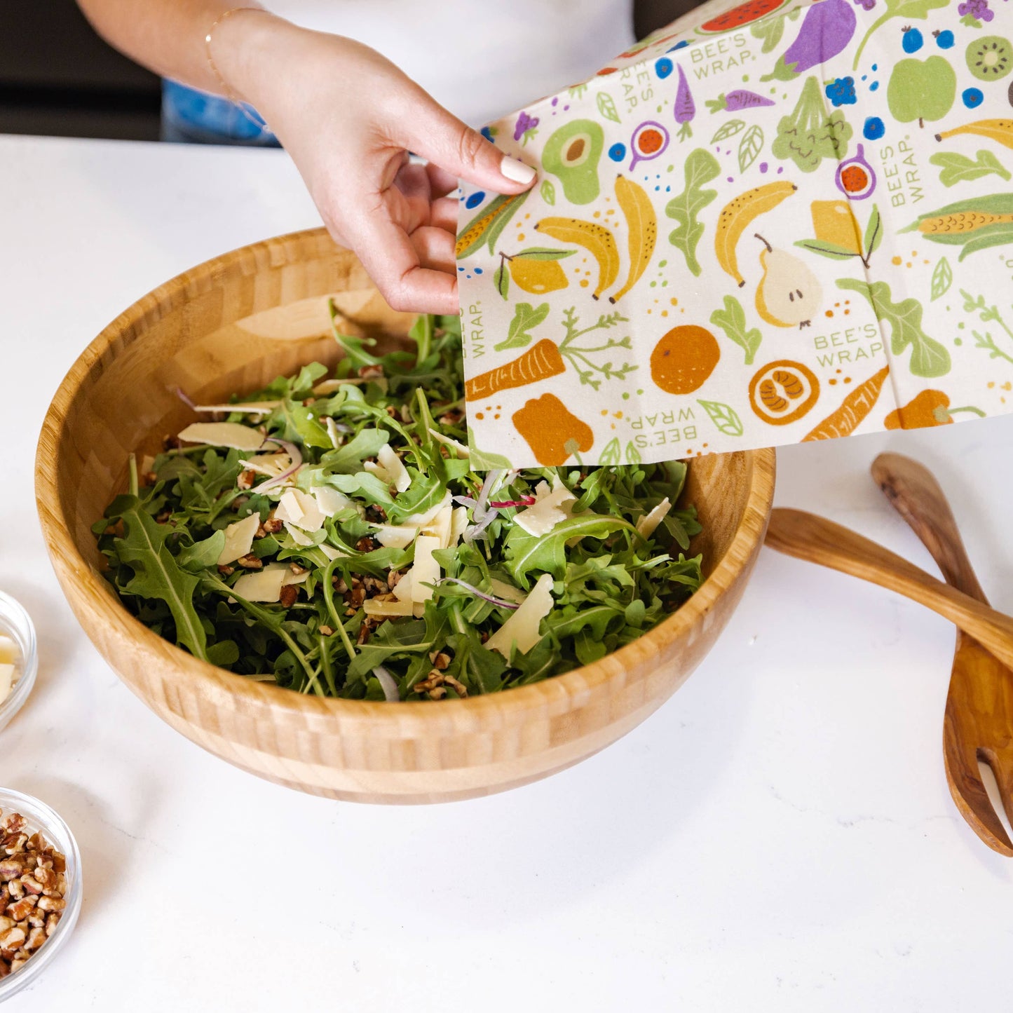 Person preparing a salad with a colorful towel featuring fruits and vegetables on a white table.