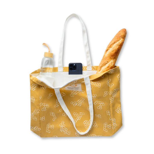 Yellow tote bag with white floral pattern containing a bottle, phone, and bread on a white background