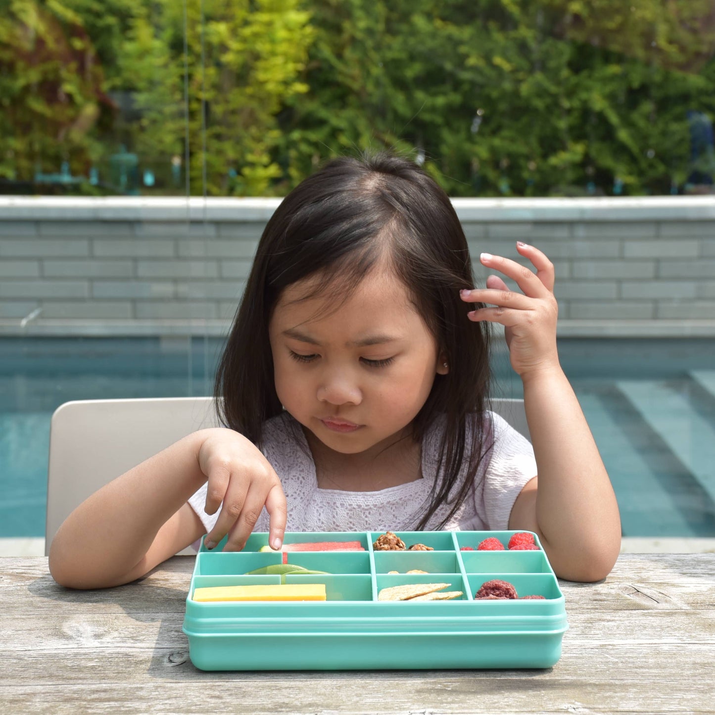 Child sitting at a table with a teal compartmentalized container outdoors near a pool.