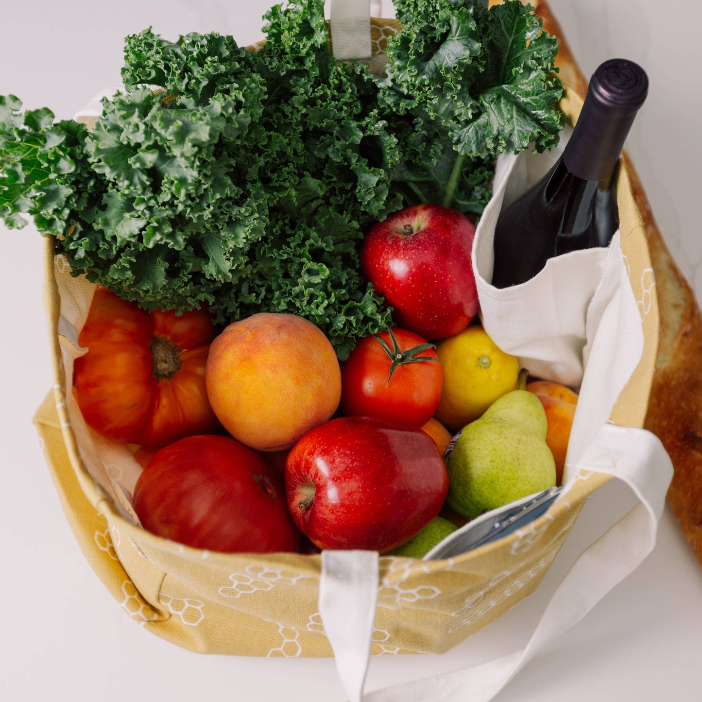 Basket filled with fruits, vegetables, and a bottle of wine on a white surface