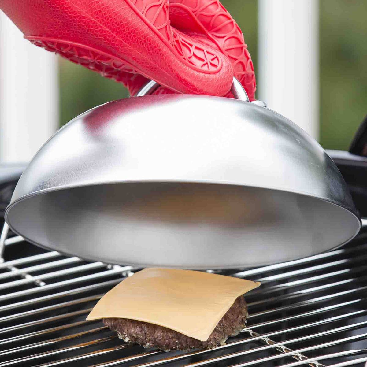 Metal dome being used to cook a burger on a grill with a red tool.