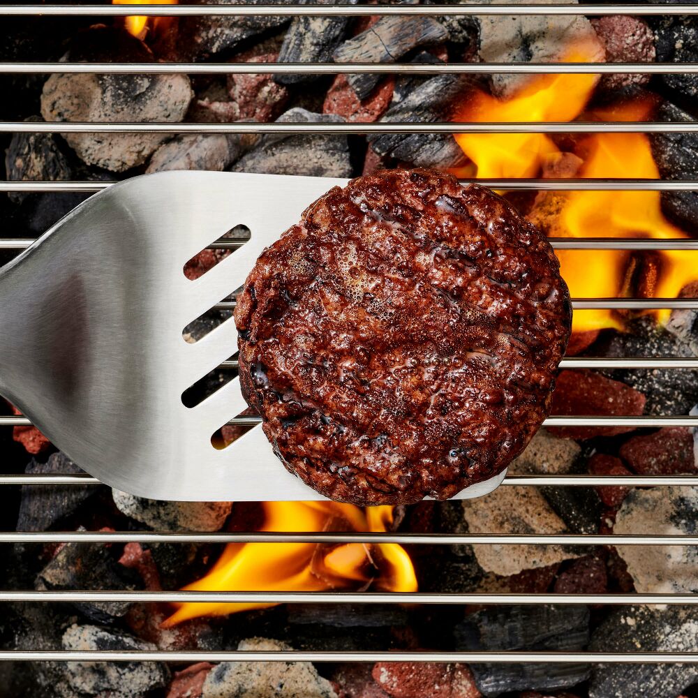 Hamburger patty being flipped on a grill with flames and coals visible