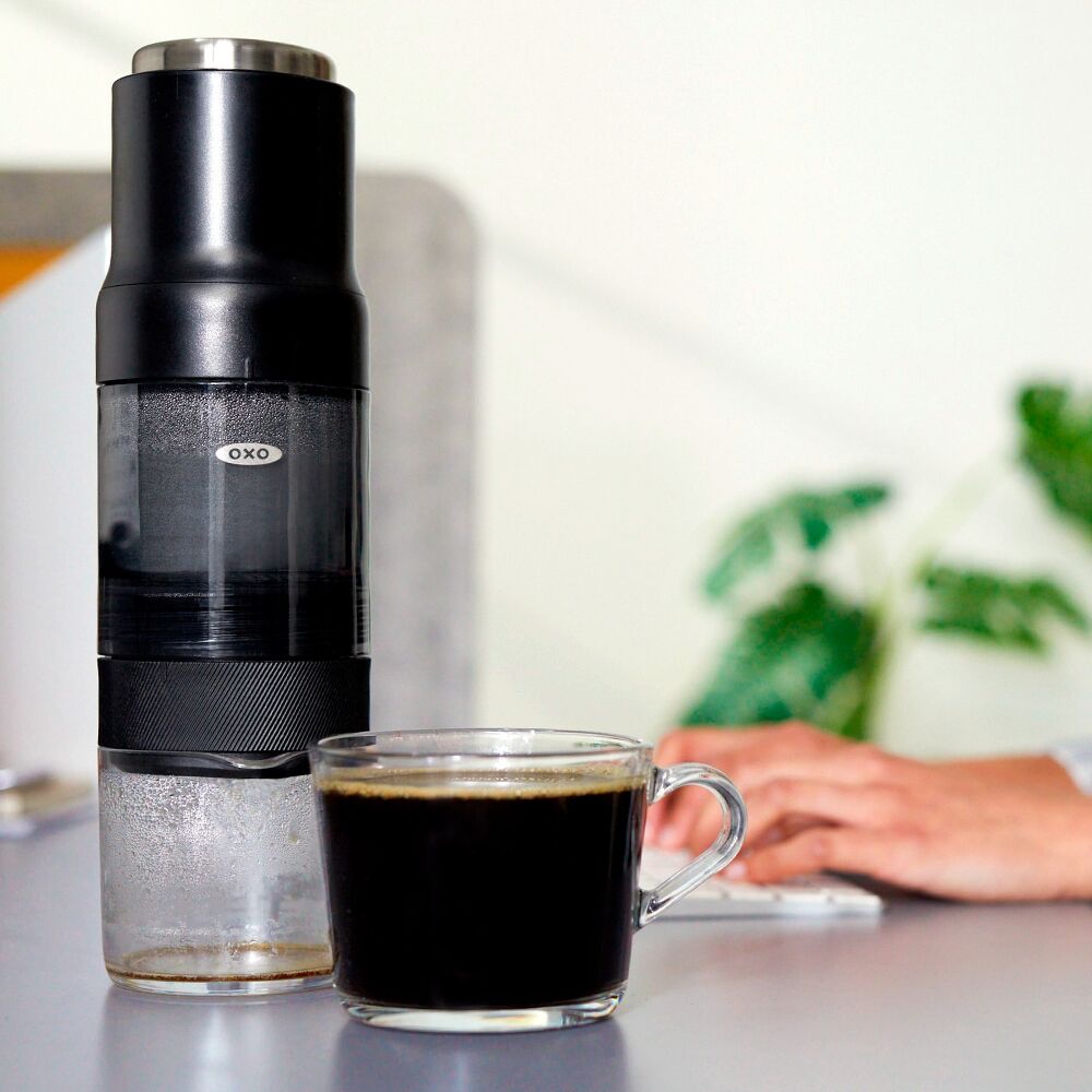 Black OXO coffee maker next to a glass of black coffee on a blurred background