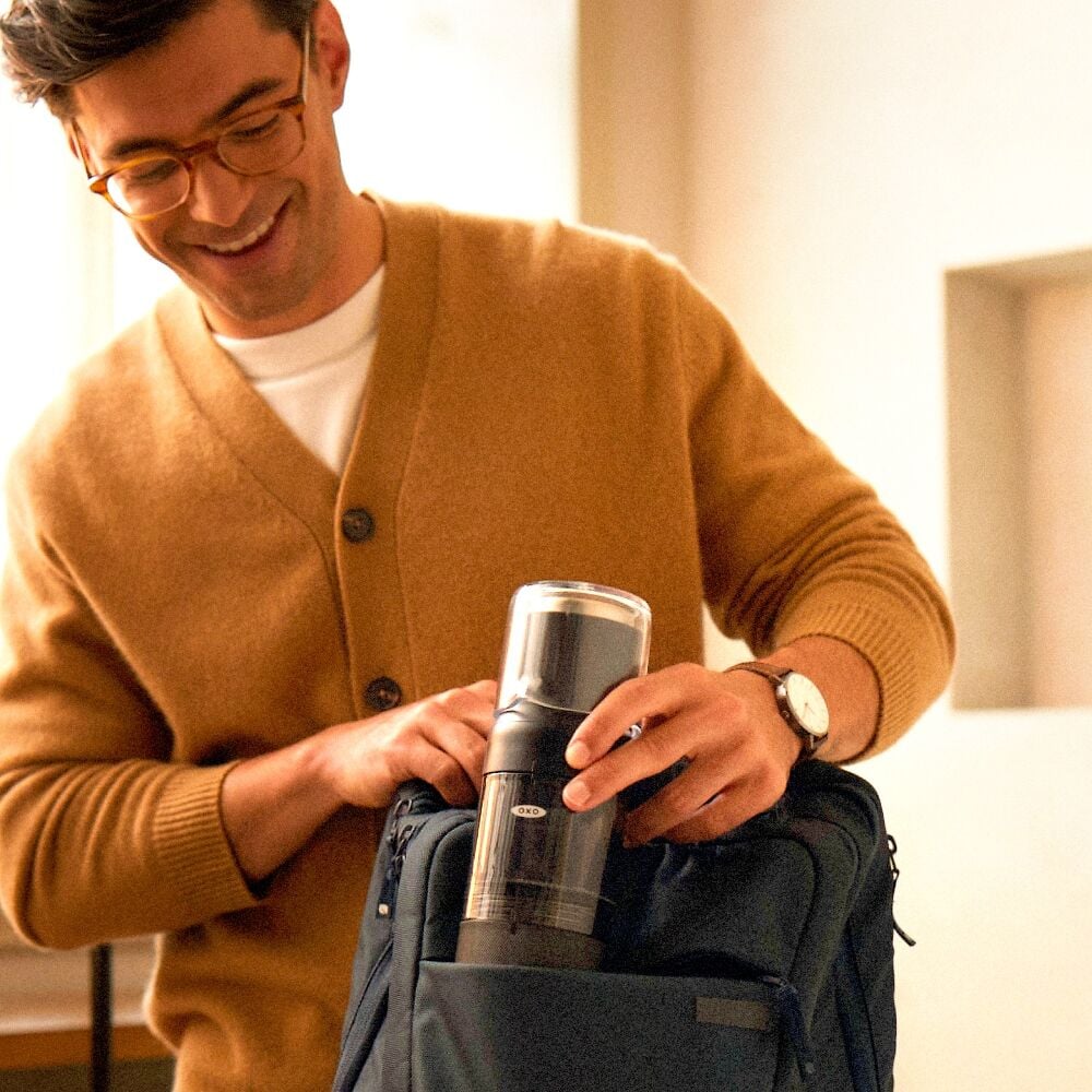 Man in a brown cardigan holding a black coffe brewer placing it into a backpack, standing indoors.