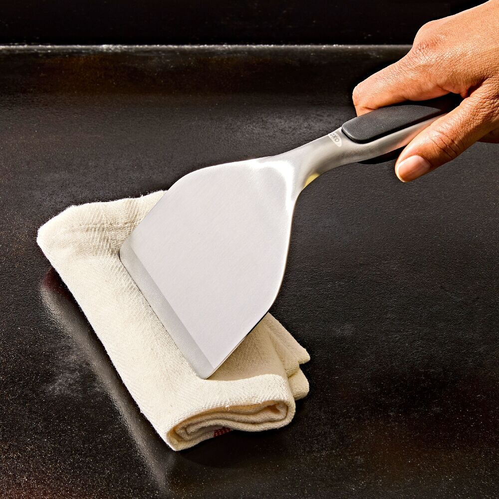 Person using a grill scraper with white towel with a black handle on a dark grill.