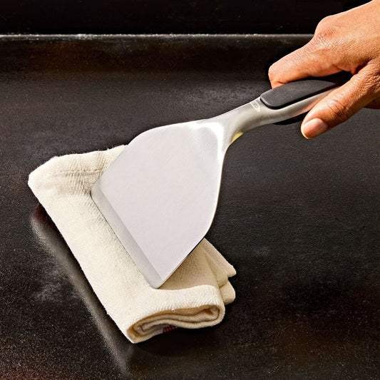 Person using a grill scraper with white towel with a black handle on a dark grill.