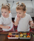 Two young girls sitting at a table with divided snack containers filled with various food items.